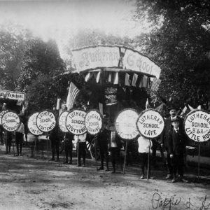 Lutheran School parade along wooded road with participants holding round signs representing various school cities, photograph hand-signed "Pappe and Doepke"