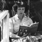 White woman seated reading on porch in blouse and skirt