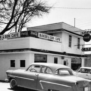 Wood frame building with signs "Baker & Son Cafe," "Coca-Cola,"  "Cafe," and "Chicken" and cars