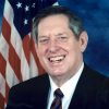 white man in suit and tie standing before American flag