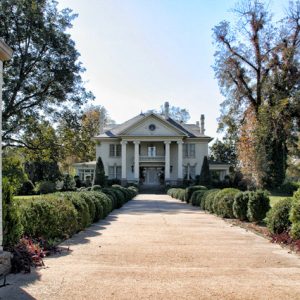 Multistory house with covered porch on walkway lined with trees and decorative bushes with brick entrance columns