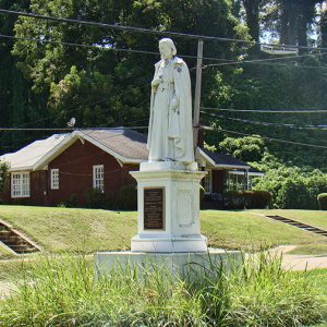 Statue of man in robes standing on a pedestal with plaque on street corner in residential neighborhood