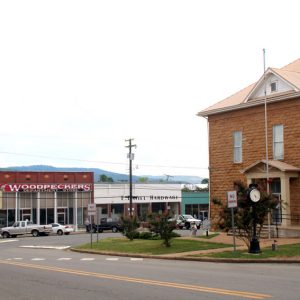 Multistory brick building with monuments flag poles and clock on two-lane road across from brick storefronts