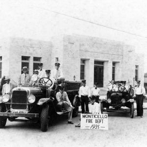 Group portrait of firemen with fire truck and vehicle with sign "Monticello Fire Dept 1935" outside brick building