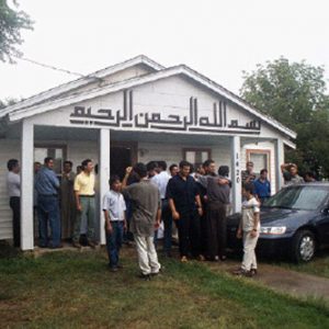 Crowd men two boys outside wood frame house under porch awning with sign featuring Arabic lettering