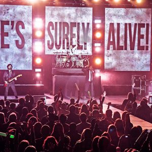 Musicians performing on stage to crowd with "He's Surely Alive" background behind them