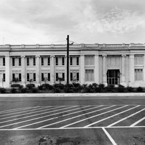 Two story classical architecture beyond sidewalk with bushes power poles and stripe painted pavement