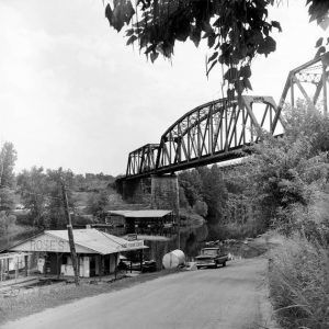 Truck parked on dirt road at "Rose's" store building next to river under steel truss bridges across from boat dock