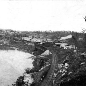 Riverside town view from wooded ridge along railroad tracks with bridge and curving shoreline