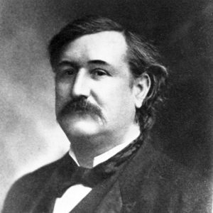 Portrait photo of a white man with a dark mustache brushed hair solemn expression in suit and bow tie