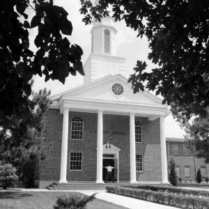 man standing with his arms outstretched on covered porch of multistory brick building with steeple and four columns