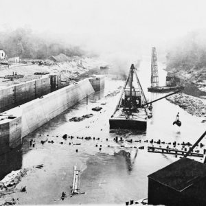 Dam construction in river with flat barge cranes, concrete lock and buildings in background