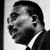 profile view of black man speaking with short hair and mustache wearing a suit and tie