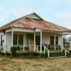 Single-story house with rusted metal roof and covered porch next to barn with water tower in the background