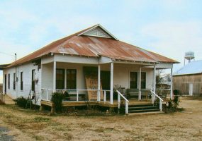Single-story house with rusted metal roof and covered porch next to barn with water tower in the background