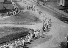 Aerial view of a parade led by school children passing through town by homes over railroad tracks