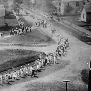 Aerial view of a parade led by school children passing through town by homes over railroad tracks