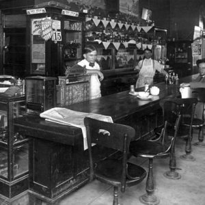 Cafe interior with two waiters, men at bar, cigar case, side doorway marked "Barber Shop"