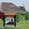 Wooden sign with Native American man on it with a brick building with a wooden roof behind it and flag pole