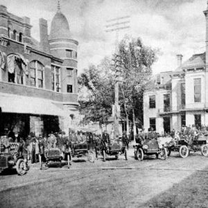 Groups of pedestrians and some cars between a multistory Victorian building and a multistory building with clock tower