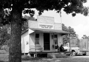 Two men seated on porch of small wood frame building under awning near truck