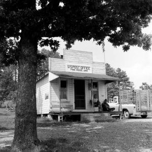 Two men seated on porch of small wood frame building under awning near truck