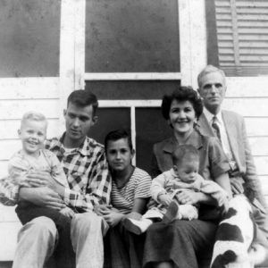 White family with 2 men, 1 woman, 3 children, and dog outside wood frame house