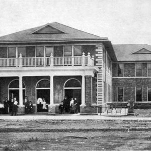 Two story brick building with arched doorways, balcony, various people, wooden cart