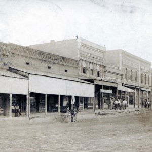 Street scene with men below awnings, carriage, wheel tracks in dirt