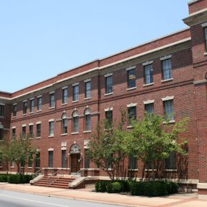 Multistory brick building with front staircase and double wooden doors bordered by hedges and small trees