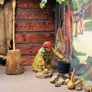 mannequin dressed as indigenous person posed with iron pot in front of mural depicting indigenous life