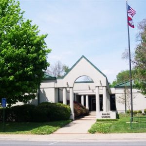 One-story white minimalist building complex with columns and a green metal roof with a walkway and bike rack and a flagpole with a US flag and Arkansas flag