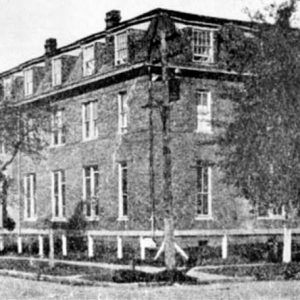 Multistory brick building viewed from street corner with surrounding fence posts and trees