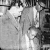 Two black men standing and one seated in library wearing suits jackets and ties gazing downwards