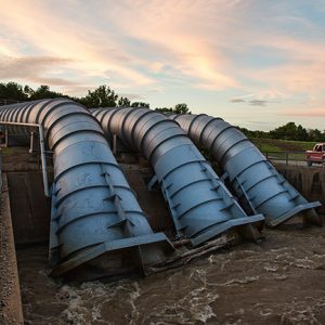 Three large pipes stretching across flat ground and acring downward into water with people watching from fence on both sides