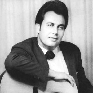 White man in suit and bow tie with dark pompadour and sideburns seated smiling with his arm on acoustic guitar