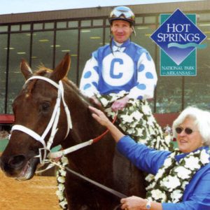 jockey sitting astride horse draped with flowers while woman the lead, with logo in top-right corner reading, "Hot Springs National Park Arkansas," and text at bottom reading, Smarty Jones, Oaklawn"