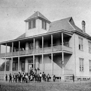 Children in yard outside large two story building with two women on balcony