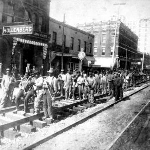 Black railroad workers stand on tracks downtown in perpendicular groups of four holding picks and hammers