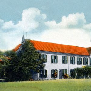 Two-story monastery with green shutters and an orange roof surrounded by trees in a grass field.