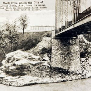 Underside of steel truss bridge over river with brick supporting columns and multistory buildings in the background with writing saying "rock from which the city of little rock Arkansas took its name"