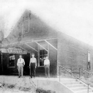 Three men in dress shirts stand on sidewalk below awning with "Tailors" sign near stairs