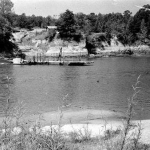 Ferry traveling down river viewed from grassy river bank with opposite bluff road and building