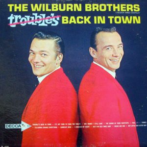 Two white men in red jackets on blue background with yellow and light blue text showing the word "trouble's" crossed out