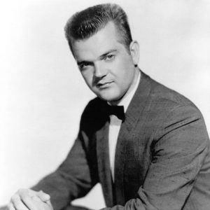 White man seated with hands crossed and a small smile with short hair combed upwards wearing suit jacket and bow tie