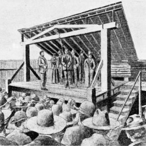 Crowd of people gathered around a gallows where white men in suits are hanging a man