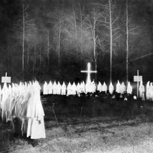 Men in white robes with pointed masks burning a cross in a forest clearing