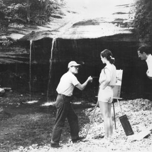 White man in cap and jeans talking to white man and woman while painting near a natural waterfall