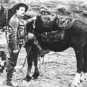 White man with mustache and cowboy hat mounting his horse while his dog stands nearby