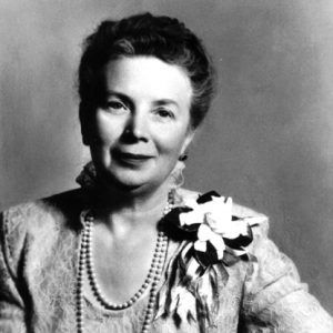 Portrait of woman with small smile and pulled back hair and hands crossed in dress pearls earrings and corsage